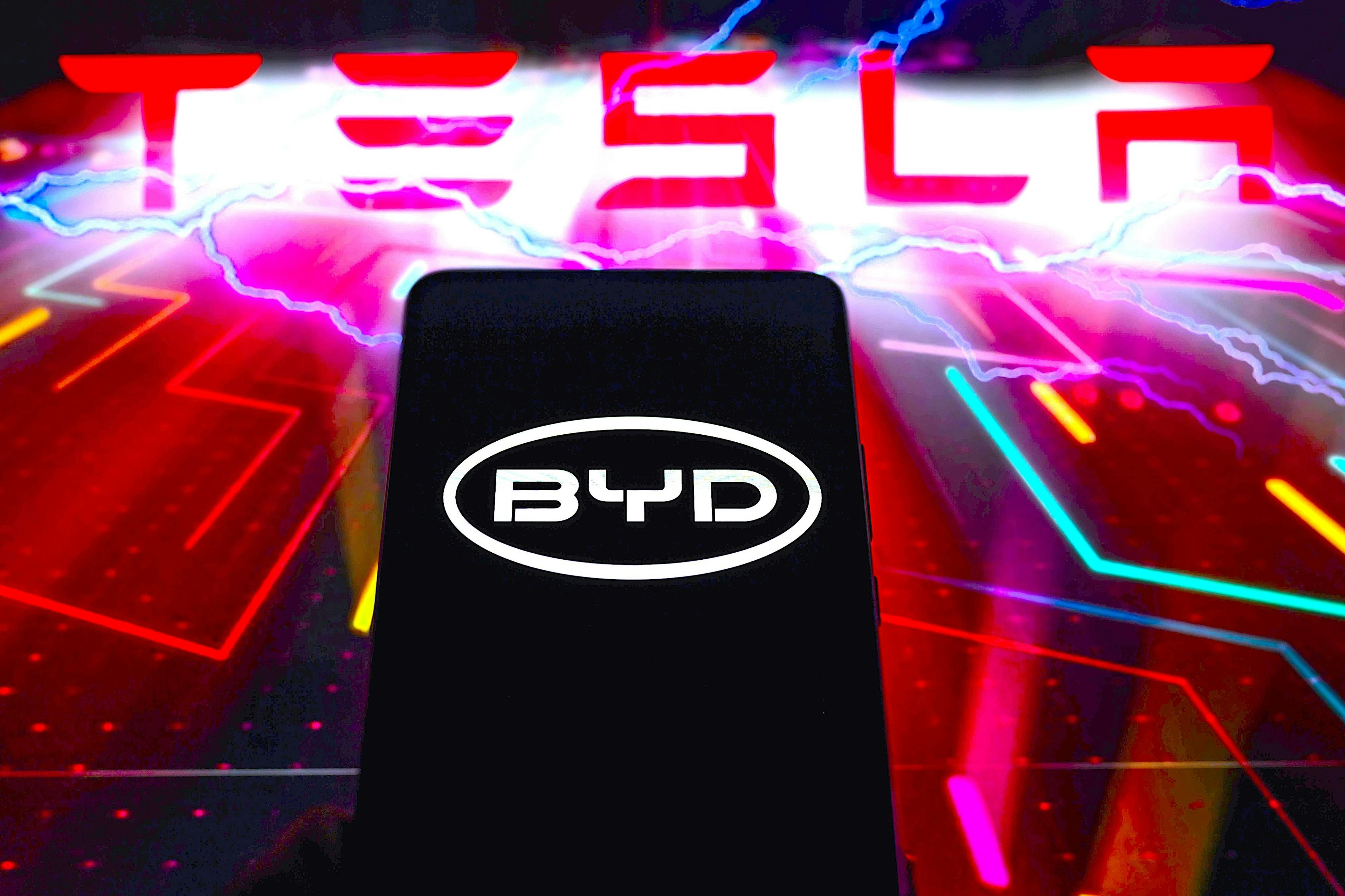 Das BYD-Logo wird auf einem Smartphone angezeigt, während im Hintergrund Tesla zu sehen ist.