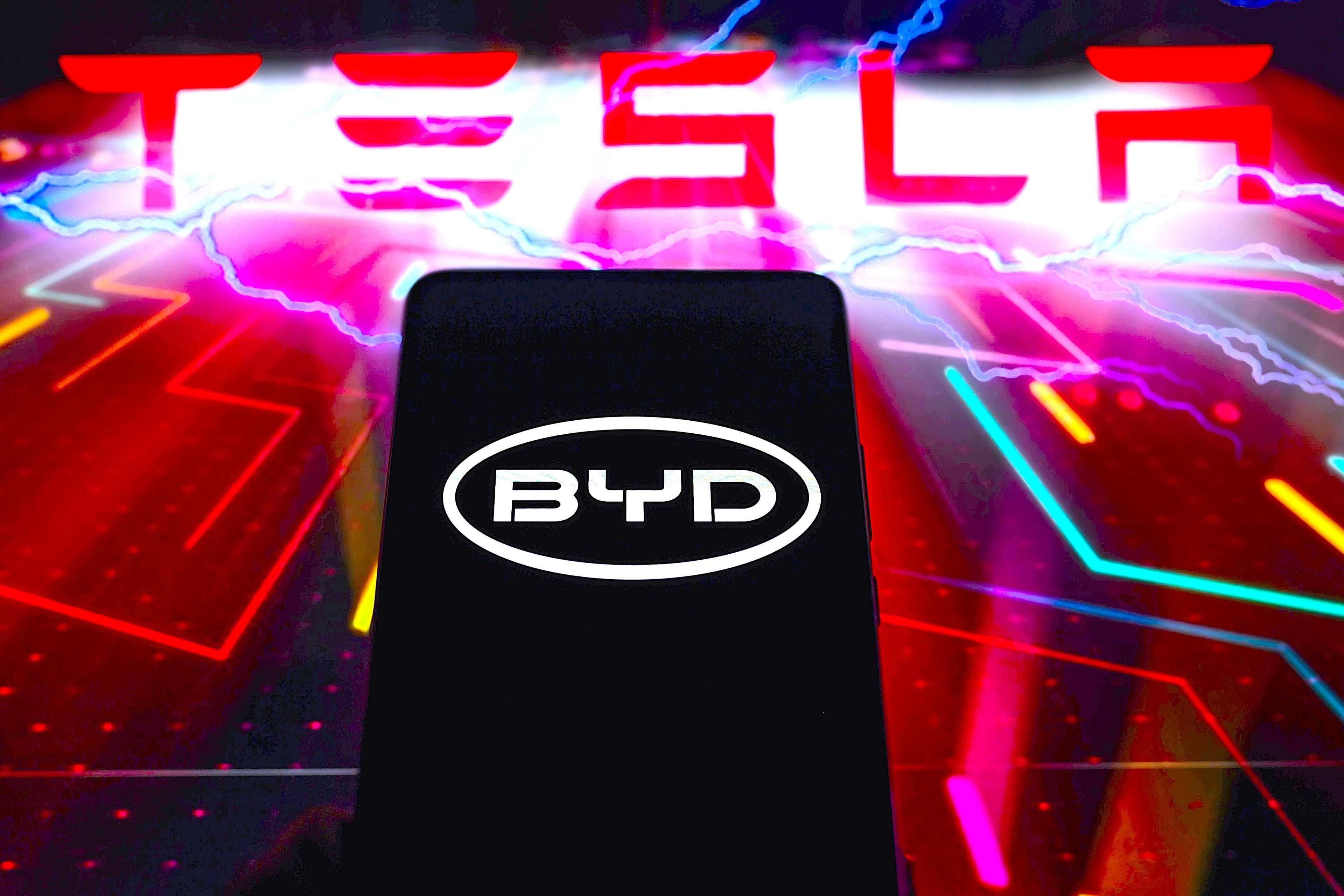 Das BYD-Logo wird auf einem Smartphone angezeigt, während im Hintergrund Tesla zu sehen ist.
