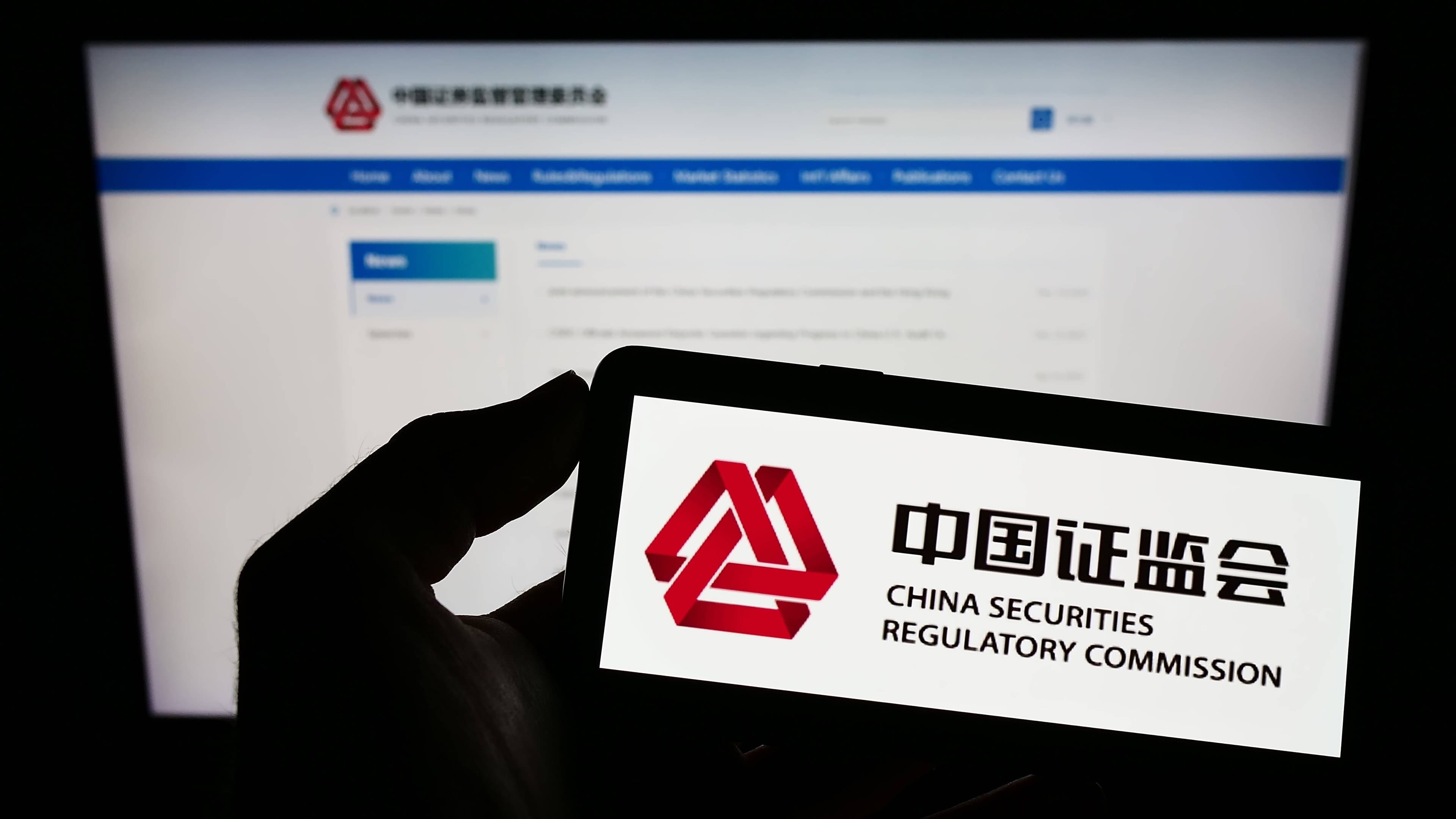 Eine Person hält ein Mobiltelefon mit dem Logo der chinesischen Wertpapieraufsichtsbehörde (CSRC) vor einem Bildschirm mit der entsprechenden Website vor sich